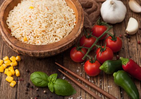Wooden bowl with boiled long grain basmati rice with vegetables on wooden table background with sticks and tomatoes with corn,garlic and basil.の写真素材