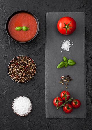 Black bowl plate of creamy tomato soup on black background with stone chopping board and raw tomatoes, pepper and salt. Top view.の写真素材