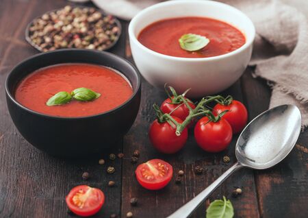 Ceramic bowl plates of creamy tomato soup with spoon, pepper and kitchen cloth on wooden board with raw tomatoes.の写真素材