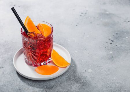 Negroni cocktail in crystal glass in white ceramic plate with orange slice and black straw on light...