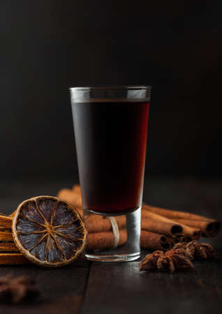Shot glass of herb liqueur with cinnamon and dry oranges on dark wooden background.