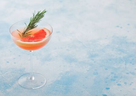 Red grapefruit summer juice cocktail in modern luxury martini glass with rosemary and fruit slice on light blue background. Space for textの写真素材