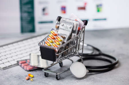 Shopping trolley with medicines for online purchase with keyboard and stethoscope on light background. Vitamins and antibiotics pills with painkillers and flu and cold capsules.の写真素材