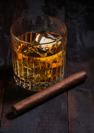 Single malt scotch whiskey in crystal glasses with ice cubes and cuban cigar on wooden table background.の写真素材