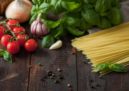 Raw spaghetti pasta in glass bowl with oil and garlic, basil and tomatoes with pepper and onion on wooden table. Top viewの写真素材