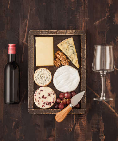 Glass and bottle of red wine with selection of various cheese in wooden box and grapes on wooden background. Blue Stilton, Red Leicester and Brie Cheese and knife.の写真素材