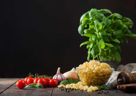 Fresh raw maccherono elbows pasta in glass bowl with basil plant, oil and tomatoes with garlic and pepper on wooden table background. Space for textの写真素材