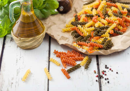 Raw homemade tricolore fusilli pasta in brown paper on white wooden table background with basil and oil.の写真素材