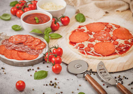 Preparation for baking of pepperoni pizza with salami spicy chorizo with wheel cutter and fresh tomatoes and basil on light table with bowl plates with cheese and tomato paste.の写真素材