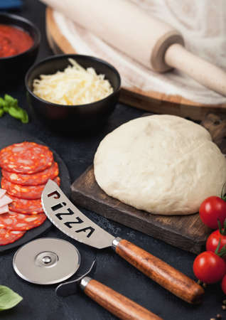 Preparation for baking of pepperoni pizza with raw dough, salami spicy chorizo with wheel cutter and fresh tomatoes and basil on dark table with bowl plates with cheese and tomato paste.の写真素材