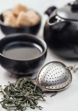 Fresh organic loose green tea with strainer infuser and black teapot and cane sugar on white table background.の写真素材