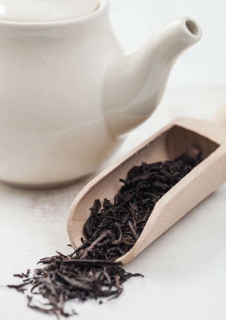 Wooden scoop of black loose tea with white ceramic teapot on white background.