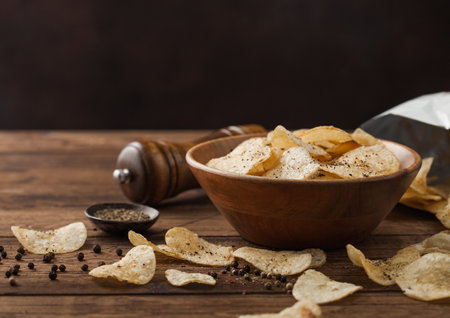 Crunchy potato crisps chips snack with black pepper in wooden bowl on dark table background with mill and ground pepper.の写真素材