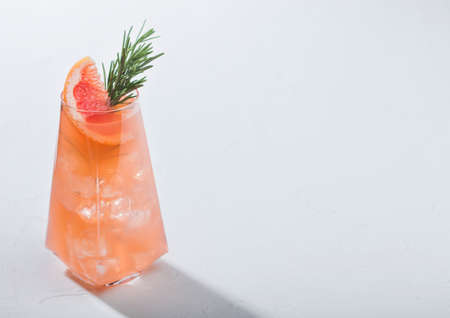 Modern luxury glass of refreshing summer red grapefruit cocktail with ice cubes, fruit slice and rosemary on white background. Space for textの写真素材