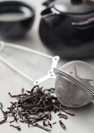 Glass jar of black loose organic tea with teapot and cup with tea strainer infuser on light background.の写真素材