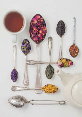 Various spoons with black and green tea, rose buds mix and blue mallow flowers and petals on white background with teapot and cup. Top viewの写真素材
