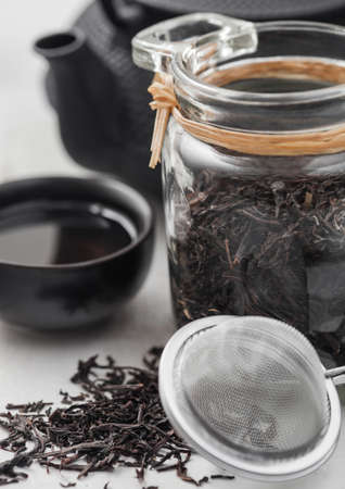 Glass jar of black loose organic tea with teapot and cup with tea strainer infuser on light background.の写真素材