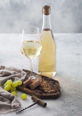 Glass and bottle of white homemade wine with corks, corkscrew and grapes on wooden board with linen cloth on light table background.の写真素材
