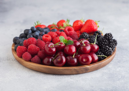 Fresh organic summer berries mix in round wooden tray on light kitchen table background. Raspberries, strawberries, blueberries, blackberries and cherries. Top viewの写真素材