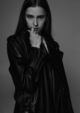 Young beautiful fashion model in long leather coat and dark jeans on dark grey.の写真素材