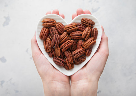 Female hands holding heart shaped plate with peeled pecan nuts on light background.