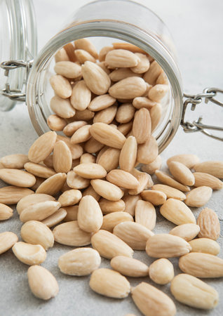 Raw white peeled almond nuts snack in glass jar on white kitchen table.