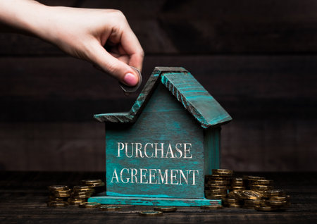 Wooden house model with coins next to it and hand holding the coin with conceptual text. Purchase Agreementの写真素材