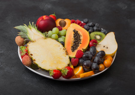 Fresh raw organic summer berries and exotic fruits in white plate on black background. Pineapple, papaya, grapes, nectarine, orange, apricot, kiwi, pear, lychees, cherry and physalis. Top viewの写真素材