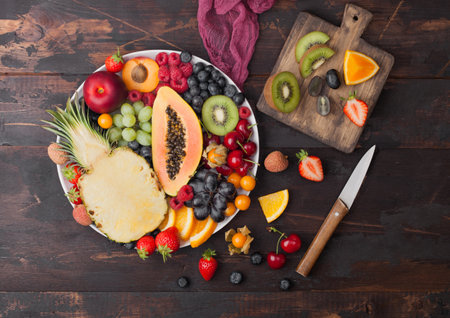 Fresh raw organic summer berries and exotic fruits in white plate on dark wooden background with chopping board and knife. Pineapple, papaya, grapes, nectarine, orange, apricot, kiwi, pear. Top viewの写真素材