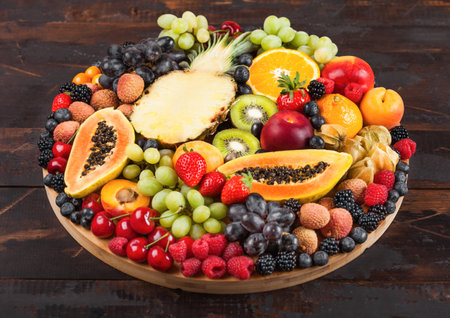 Fresh raw organic summer berries and exotic fruits in round large tray on dark wooden kitchen background. Papaya, grapes, nectarine, orange, raspberry, kiwi, strawberry, lychees, cherry. Top viewの写真素材