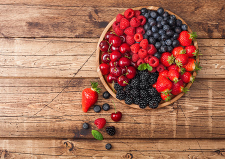 Fresh organic summer berries mix in round wooden tray on light wooden table background. Raspberries, strawberries, blueberries, blackberries and cherries. Top viewの写真素材