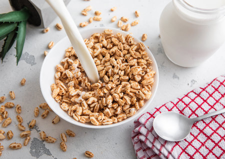 Pouring milk in to bowl with honey rice krispies morning cereal breakfast with spoon and kitchen towel.の写真素材
