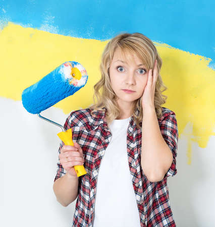 Tired woman at work in the home - painting wall at room. Portrait of woman painting big Ukrainian flag on wall at home.の写真素材