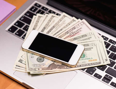 Workplace with money and electronic devices - cellphone and laptop computer. Mobile phone and US dollar banknotes on keyboard of notebook. Concept of payment and savings.の写真素材