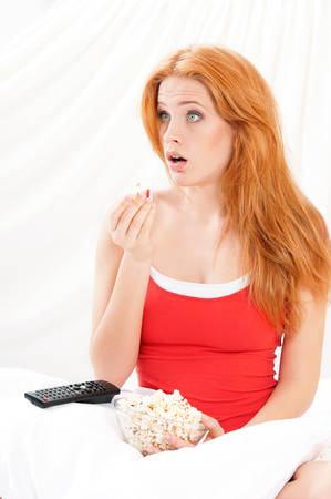 Portrait of surprised beautiful girl open-mouthed watching movie or tv in bed and eating popcornの写真素材