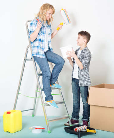 Happy mother and son makes repairs at home. Smiling beautiful young woman with teen boy painting wall at room.の写真素材
