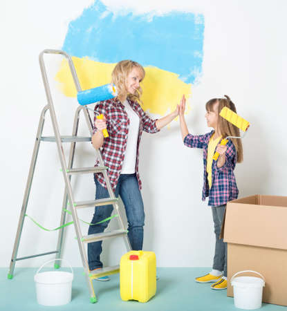Happy mother and daughter makes repairs at home - painting big Ukrainian flag on wall at room.の写真素材