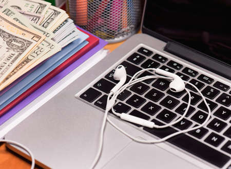 Keyboard of open laptop computer with headphones for music and money US dollar banknotes on a wooden table. Workplace with notebook.の写真素材