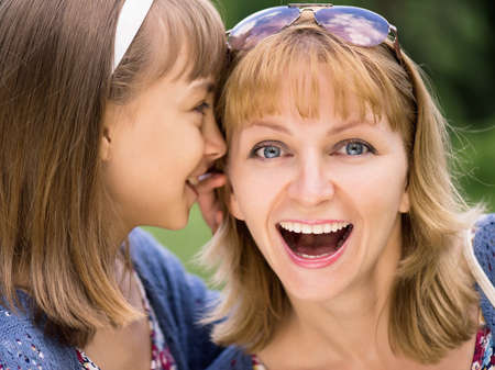 Smiling mother and daughter whispering gossip - relationships and happiness concept. A little girl telling her mother a secret. Portrait of happy family - sharing secrets, outdoors at park.の写真素材