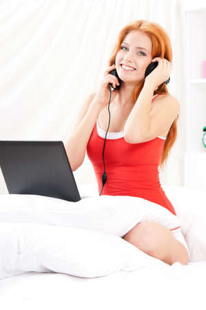Beautiful happy young woman with headphones and laptop watching a movie or working on notebook and listening to music in bed. Girl sitting on bed surfing internet or watching media.の写真素材