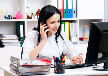 Smiling medical nurse with telephone and laptop. Portrait of happy young doctor talking on phone at office. Young woman doctor sitting with mobile and stethoscope.の写真素材