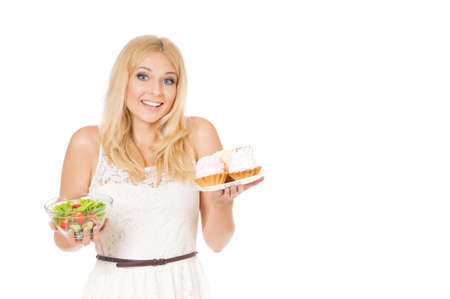 Half-length portrait of very beautiful woman holding small cake, fresh vegetables. Young housewife choosing sweets or healthy eating - cake and salad. Isolated on white background.の写真素材