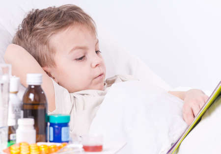Sick child boy lying in bed with fever, resting at home. Illness kid reading book.の写真素材