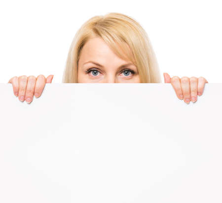 Young woman with surprised eyes peeking out from behind billboard paper poster. Businesswoman holding big white banner, isolated on white background.の写真素材