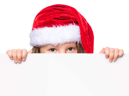 Little girl wearing Santa Claus hat, posing behind white panel isolated on white background. Kid holding empty Christmas billboard. Child peeping behind blank holidays board.の写真素材