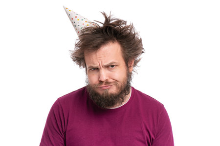 Crazy bearded Man with funny Haircut in birthday cap, isolated on white background. Unappy guy makes...