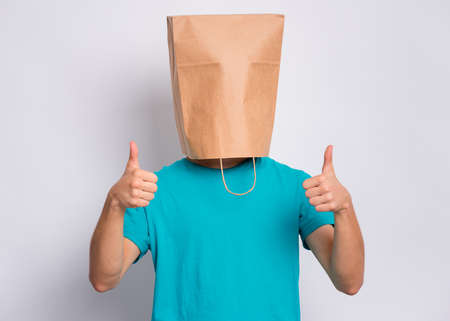 Portrait of teen boy with paper bag over head making thumb up gesture. Teenager cover head with bag showing success sign posing in studio. Child pulling paper bag over head.の写真素材