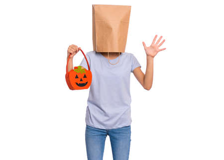 Happy halloween concept. Teen girl with paper bag over her head holds orange pumpkin bucket, isolated on white background. Portrait of teenager cover head with bag. Child waiting candy.の写真素材