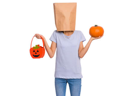 Happy halloween concept. Teen girl with paper bag over her head holds orange pumpkin bucket and small pumpkin, isolated on white background. Teenager cover head with bag. Child waiting candy.の写真素材