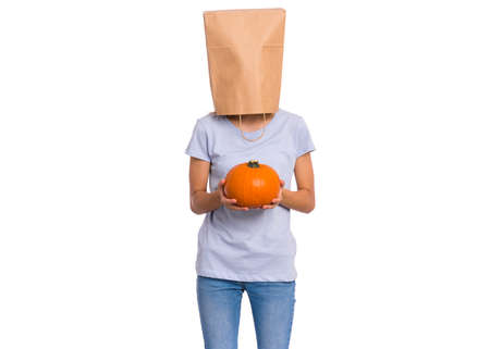 Happy halloween concept. Teen girl with paper bag over her head holds orange small fresh pumpkin, isolated on white background. Teenager cover head with bag. Child with food - raw vegetables in hands.の写真素材